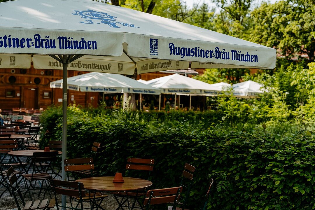 Ein großer Sonneschirm mit dem blauen Augustiner Aufdruck ist im Fokus. Darunter sieht man ein paar Tische und Stühle die im sonnigen Biergarten stehen.
