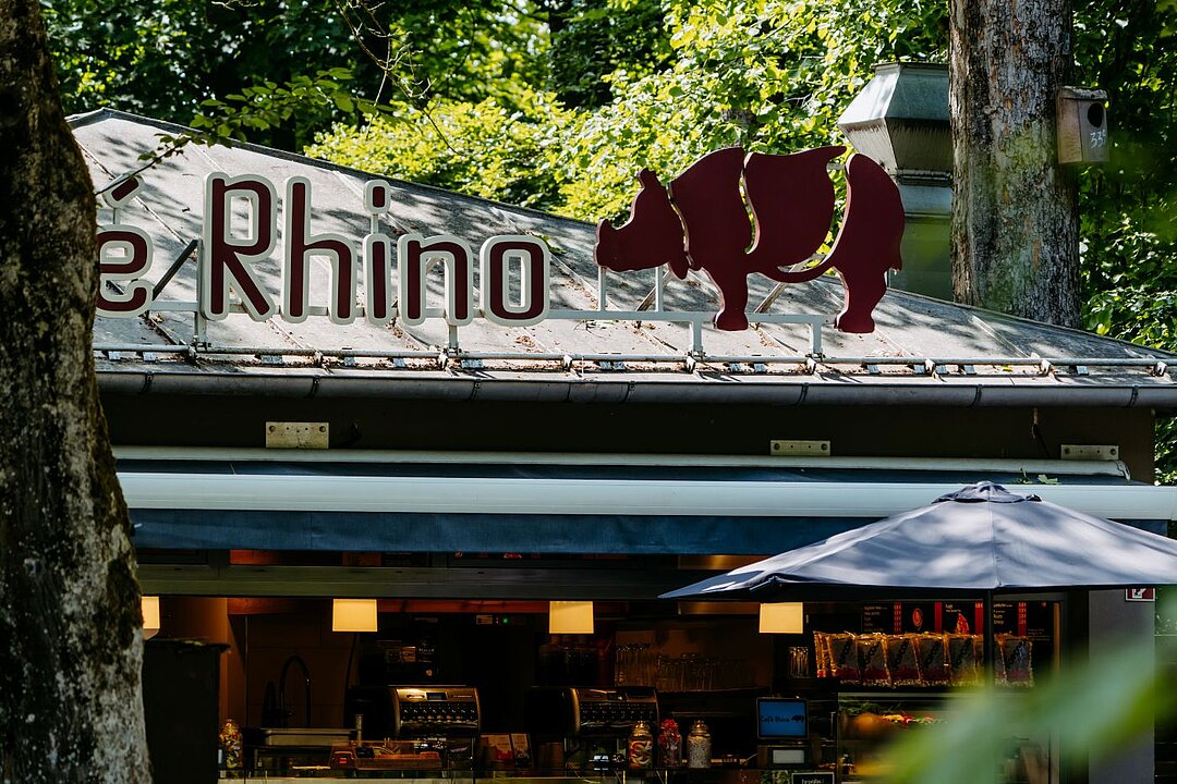 Auf dem Kioskdach sind große rot-weiße Buchstaben zu sehen, die das Wort Rhino zeigen. Daneben ist in der selben Farbe ein Nashorn zusehen.