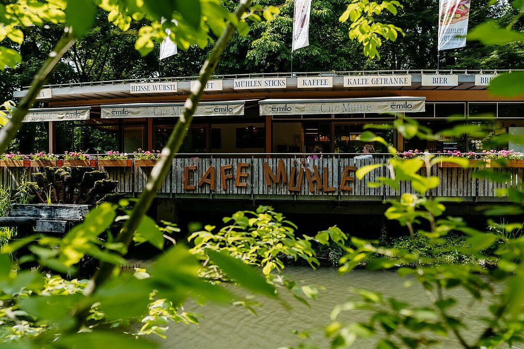 Im unscharfen Vordergrund sind saftig grüne Äste. Durch einen kleinen Fluss davon getrennt steht ein kleines Café in Holzoptik.