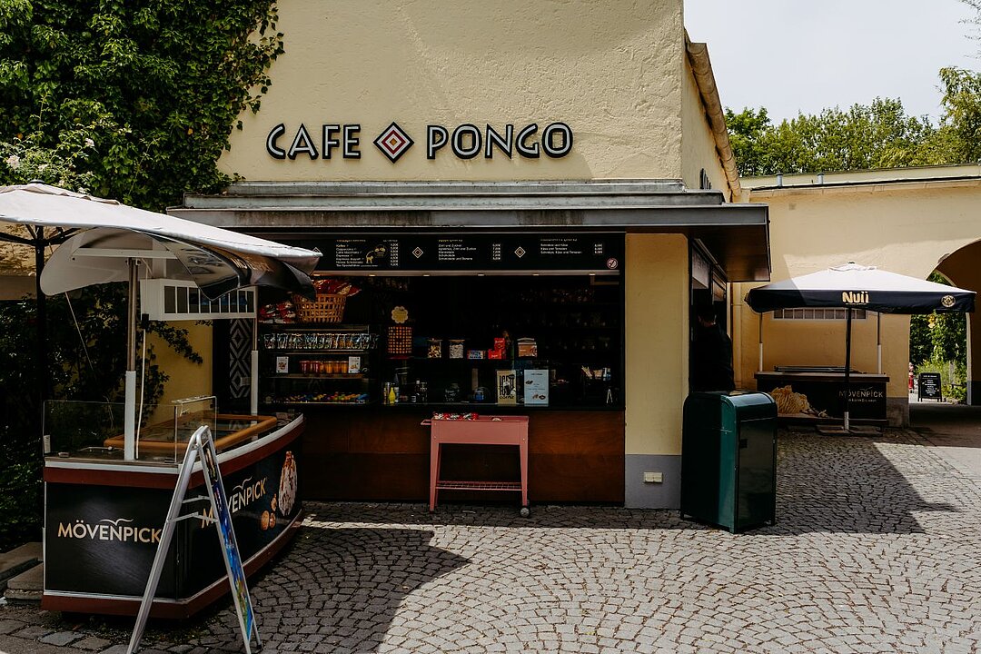 Die Sonne strahlt auf das buttergelbe Gebäude. Vorne Links und hinten rechts ist jeweils ein Sonnenschirm gespannt