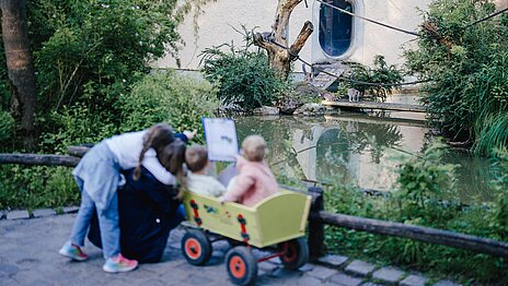 Sommer-Quizrallye im Tierpark Hellabrunn Drei Kinder sowie eine Mutter beobachten die Kattas, zwei Kinder sitzen in einem grünen Bollerwagen.